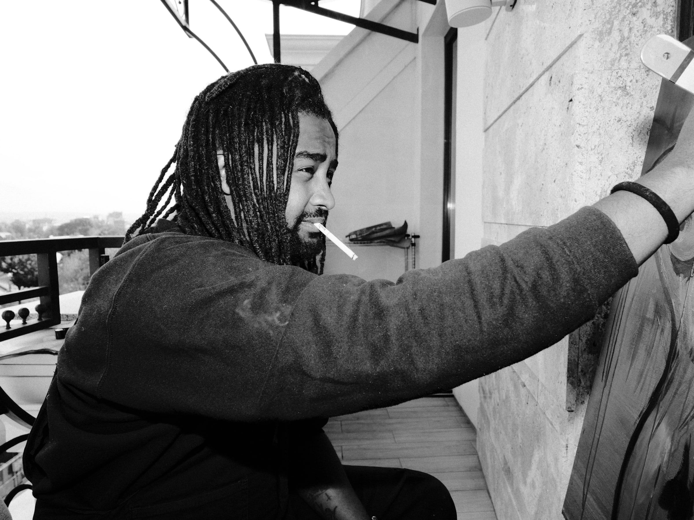 Dawit reaching up to paint a canvas propped against a wall, a cigarette in his mouth, in black and white