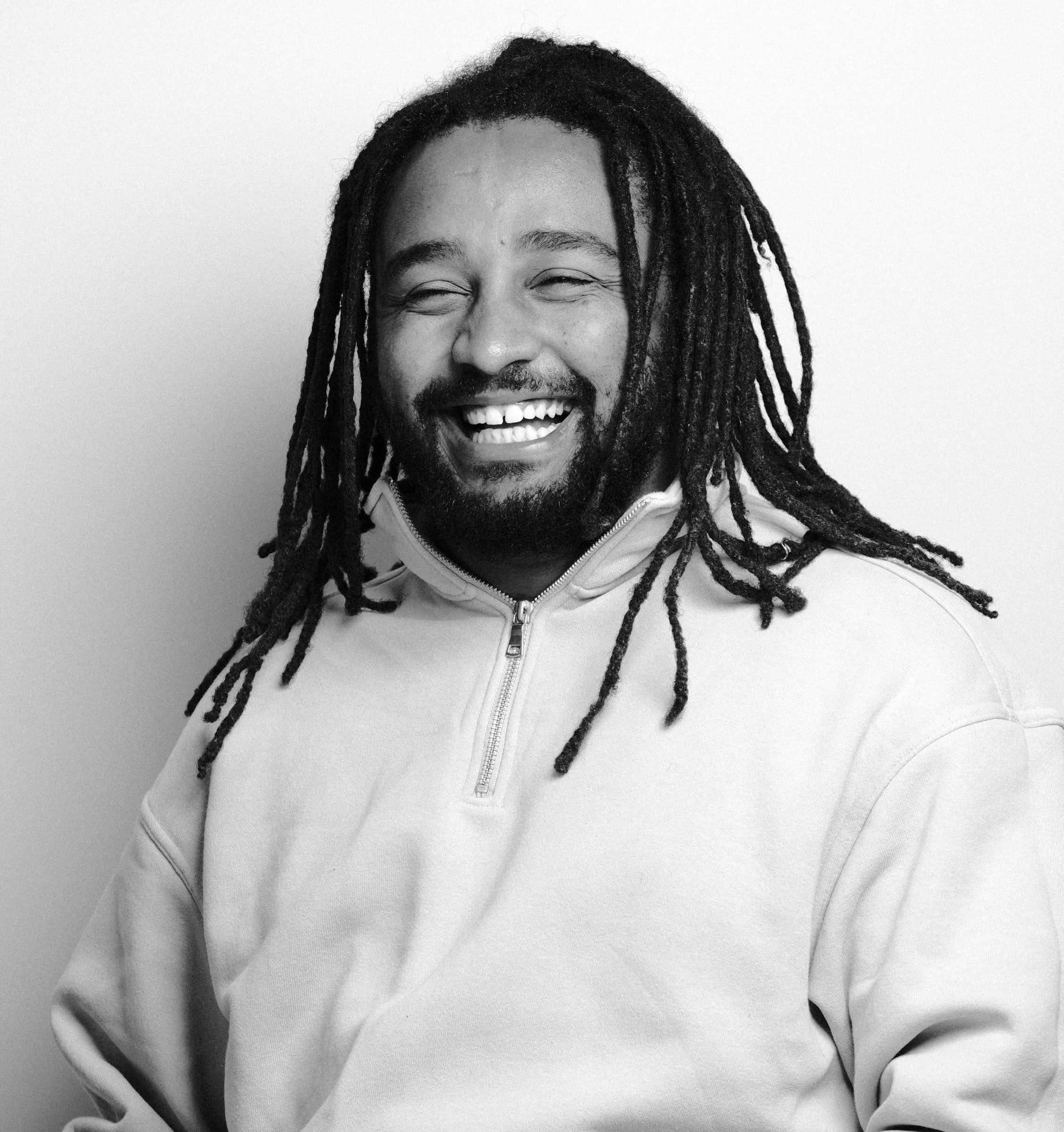 Dawit laughing in a pale quarter-zip sweatshirt against a white wall, in black and white